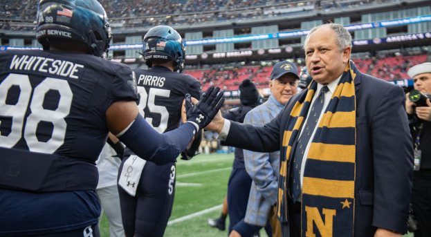 Ein Mann in Anzug und Krawatte gibt einem Fußballspieler die Hand auf einem Feld, umgeben von einer Gruppe von Menschen, mit einer Person, die eine Kamera auf der rechten Seite hält und einem Stadion voller Menschen im Hintergrund, mit Schildern, die "Notre Dame Football Coach Mike McCarthy und die Fighting Irish" lesen.