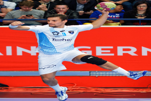 Ein Mann in der Luft mit einem Handball, mit einer Gruppe von Menschen und einem Banner mit Text im Hintergrund.