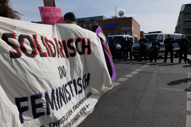 Eine Gruppe von Menschen marschiert auf einer Straße, hält ein Banner mit der Aufschrift "Solidarität und Feminismus" und hat geparkte Fahrzeuge und Gebäude im Hintergrund unter einem klaren blauen Himmel.