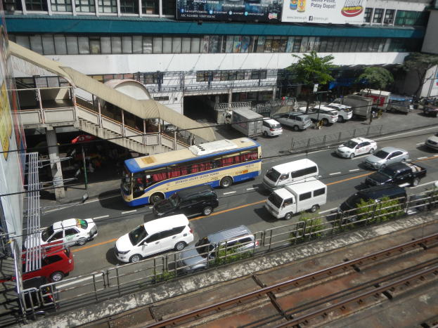 Eine belebte Stadtstraße mit Fahrzeugen, Gebäuden, Grünflächen und verschiedener städtischer Infrastruktur.