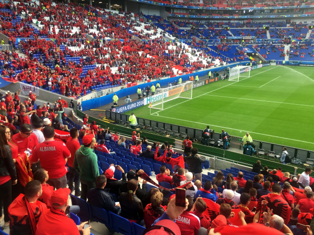 Eine große Menschenmenge in einem Stadion schaut einem Fußballspiel zu, mit Sitzplätzen und Stehplätzen, Schildern, einem Torpfosten und Deckenleuchten.
