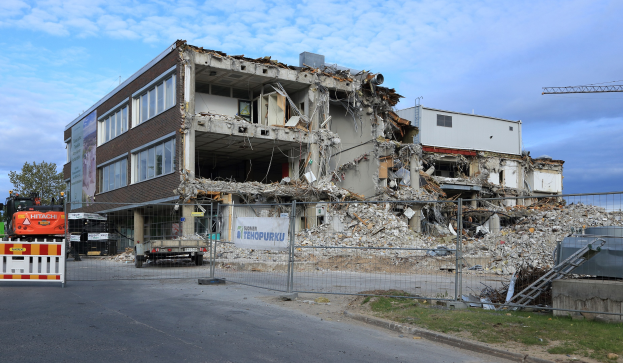 Ein großes Gebäude abgerissen in der Mitte einer Straße, umgeben von einem Metallzaun, Fahrzeugen, Gras, einem Schild, einer Fahne, Pfählen, einem Kran, Bäumen und einem bewölkten Himmel.