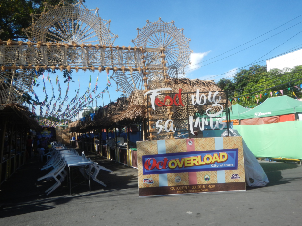 Eine Karnevalszene mit einem großen Riesenrad in der Mitte, umgeben von Hütten, Tischen, Stühlen, einem Schild, Dekorationen, Bäumen, Stromleitungen und einem bewölkten Himmel.
