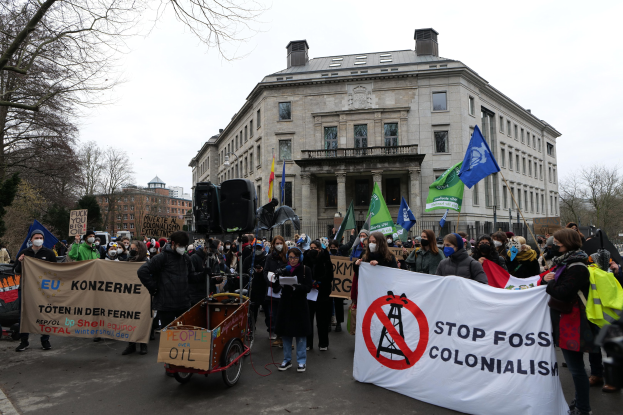 Große Gruppe von Menschen marschiert bei einer Demonstration gegen fossile Brennstoffe, trägt Transparente und Fahnen, mit einem Fahrzeug im Vordergrund und Gebäuden, Bäumen und einem klaren blauen Himmel im Hintergrund.