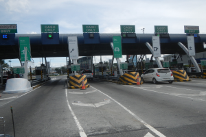 Eine Mautstation an der Seite einer Straße mit darunter fahrenden Autos, umgeben von Verkehrskegeln, Absperrpfosten, Schildern, einem Tor, Laternenmasten, Bäumen, Gebäuden und einem bewölkten Himmel.