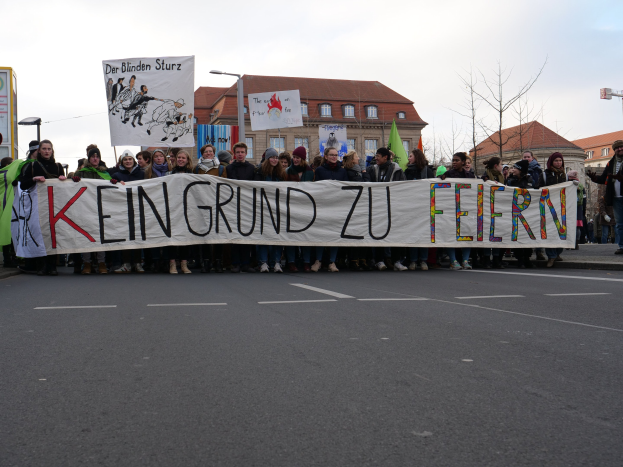 Demonstranten mit einem Banner mit der Aufschrift "Kein Grund zu Feiern" gegen deutsche Sparmaßnahmen auf einer städtischen Straße mit Gebäuden, Bäumen und einem klaren Himmel.