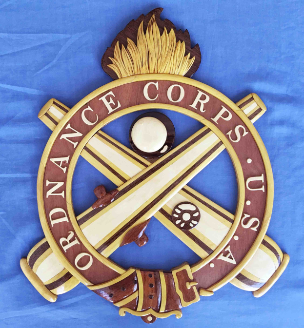 Holzschild mit "Ordnance Corps" in schwarzer Schrift, das ein weißes Stern auf einem navy-blauen Hintergrund mit einer goldenen Umrandung zeigt, auf blauem Tuch montiert.