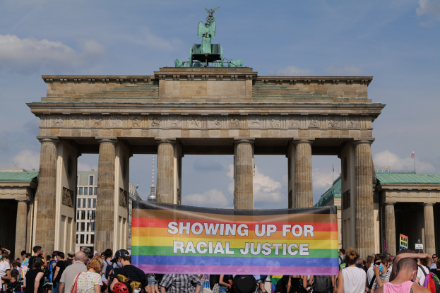 Eine Gruppe von Menschen hält eine "Racial Justice"-Schleife vor dem Brandenburger Tor in Berlin, Deutschland, mit den Säulen und der Statue des Tors im Hintergrund und Gebäuden und einem bewölkten Himmel.