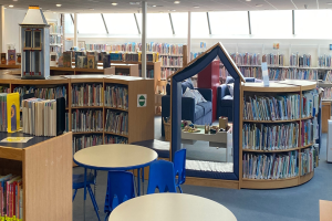 Eine Bibliothek mit Bücherregalen, Tischen, Stühlen, Sofas mit Kissen, einer Säule, Deckenleuchten und anderen Gegenständen.