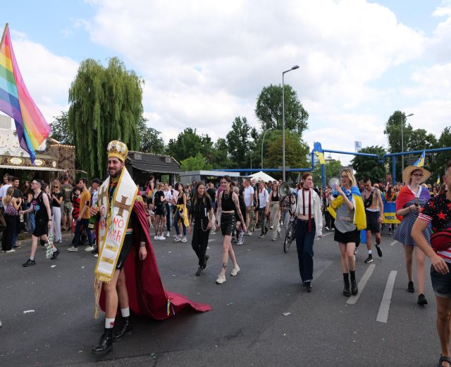 Eine Gruppe von Menschen marschiert bei der Pride Parade 2018, einige halten Musikinstrumente und tragen Mützen, eine Regenbogenfahne schwingend, mit Laternenmasten, Bäumen, Hütten und einem bewölkten Himmel im Hintergrund.