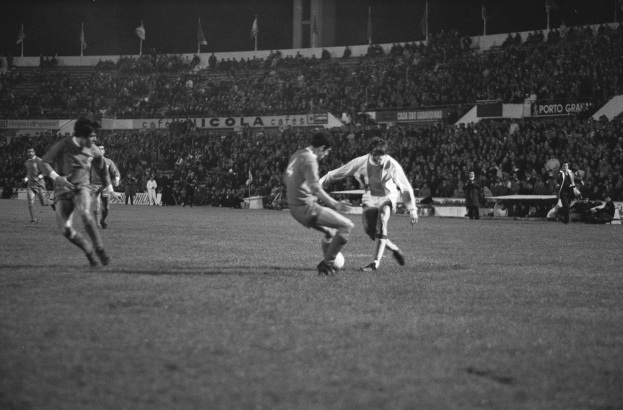 Schwarze-Weiß-Foto von Männern, die Fußball in einem Stadion mit Zuschauern und Fahnenmasten im Hintergrund spielen.
