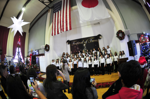 Eine Gruppe von Menschen auf einer Bühne mit Weihnachtsdekoration, ein Podium mit einem Mikrofon, eine Tafel mit Text und eine Menge mit Kameras und Handys.