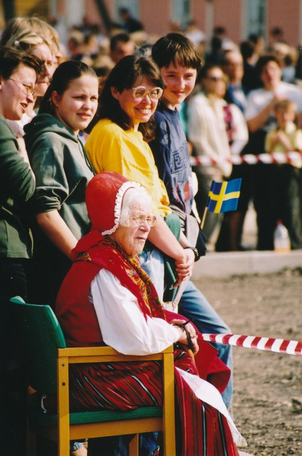 Alte Frau sitzt in einem Stuhl vor einer Menge, mit einer Schleife im Vordergrund und einem Gebäude im Hintergrund.