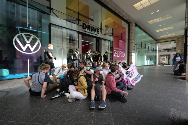 Eine Gruppe von Menschen sitzt vor einer Glasfassade eines Volkswagen-Händlers auf dem Boden und hält ein Transparent mit Text, unter Deckenleuchten, scheinbar protestierend.