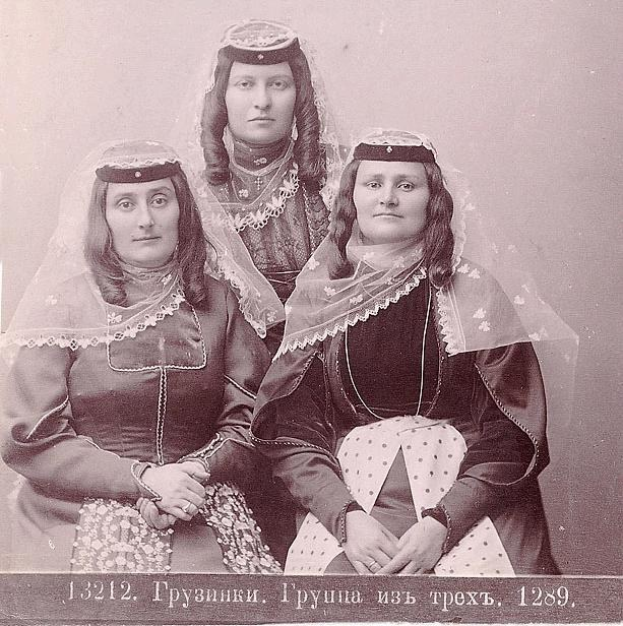 Schwarzes und weißes Foto von drei Frauen in traditionellen russischen Hochzeitskleidern mit farbenfroher Stickerei und Perlen, die vor einer weißen Wand stehen.