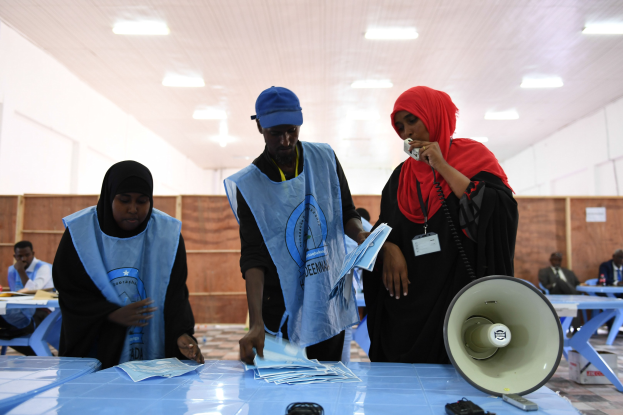 Eine Gruppe von Menschen steht um einen Tisch mit einem Megafon und Papieren, w├Ąhrend andere im Hintergrund eines Wahllokals mit beleuchteter Decke sitzen oder stehen.