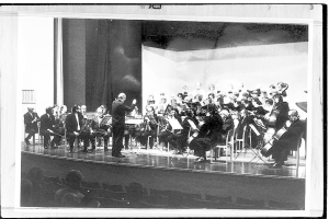 Ein Schwarz-Weiß-Foto eines Dirigenten in Anzug und Krawatte, der auf einer Bühne steht und ein Orchester mit verschiedenen Instrumenten wie Geigen, Celli und Flöten leitet, während ein Publikum auf Stühlen aufmerksam zuhört.