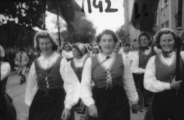 Eine Gruppe von Frauen in traditioneller bayischer Tracht spaziert strahlend durch eine Straße, umgeben von Bäumen und Gebäuden, auf einem Schwarz-Weiß-Bild.