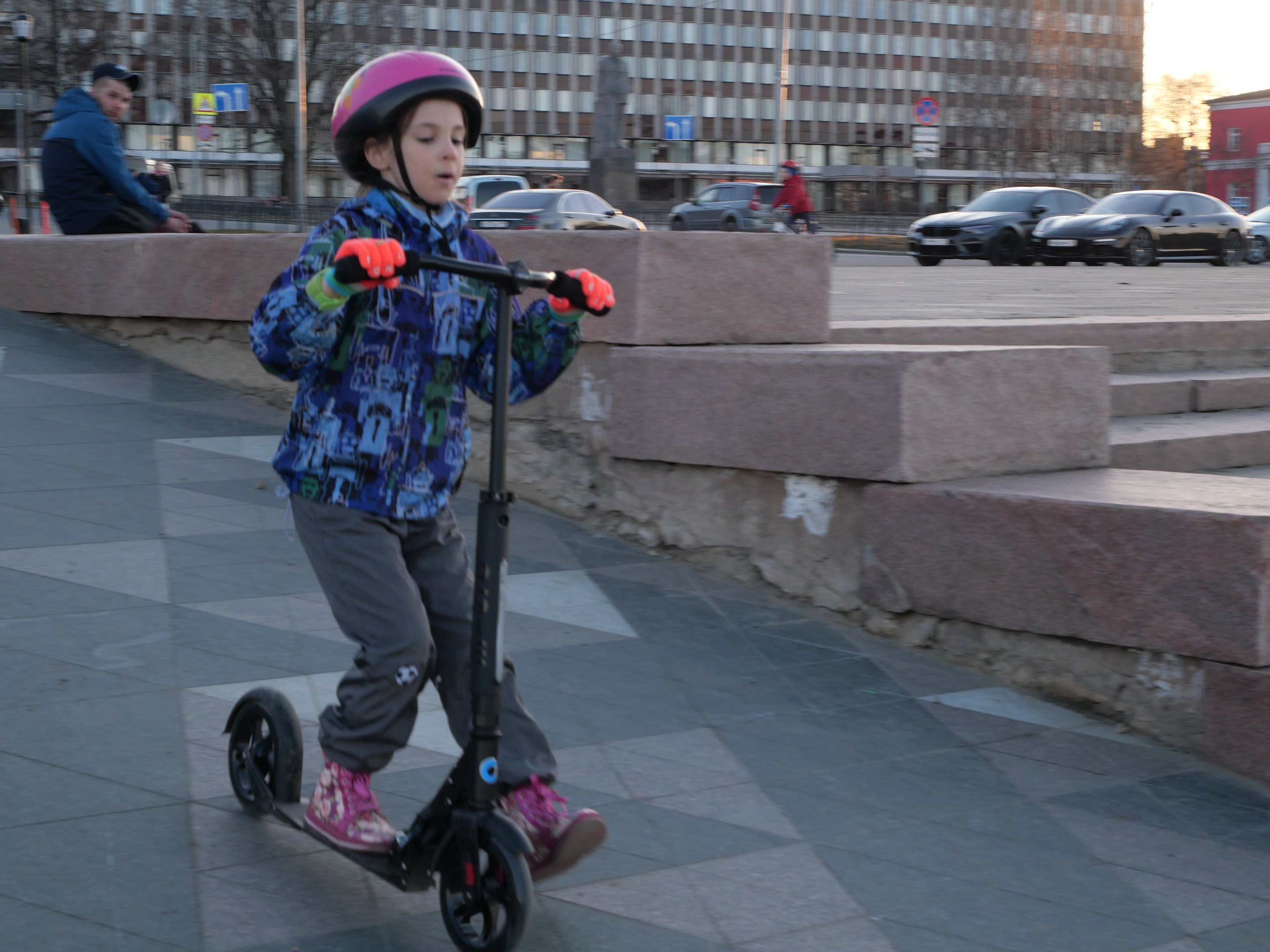 Ein junger Junge mit Helm und Handschuhen fährt auf einem Roller eine Treppe auf dem Gehweg hinunter, mit Fahrzeugen, Menschen, Bäumen, Pfählen, Brettern, Gebäuden und einem klaren blauen Himmel im Hintergrund.