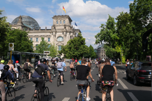 Eine Gruppe von Menschen, die Fahrräder auf einer von Bäumen gesäumten Straße in Berlin, Deutschland, fahren, mit Gebäuden auf beiden Seiten, einer Bushaltestelle auf der rechten Seite und einer Flagge, die auf einem der Gebäude weht, unter einem bewölkten Himmel.