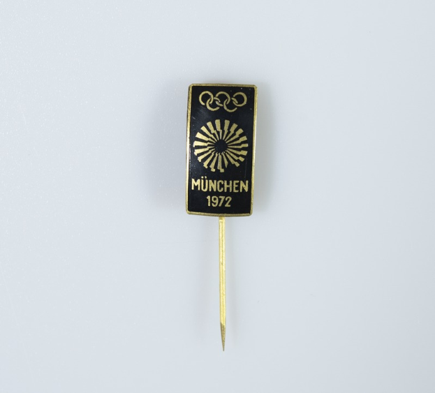 Ein schwarzer und goldener Anstecker mit der Aufschrift 'München 1972 Olympische Spiele' auf einem weißen Hintergrund.