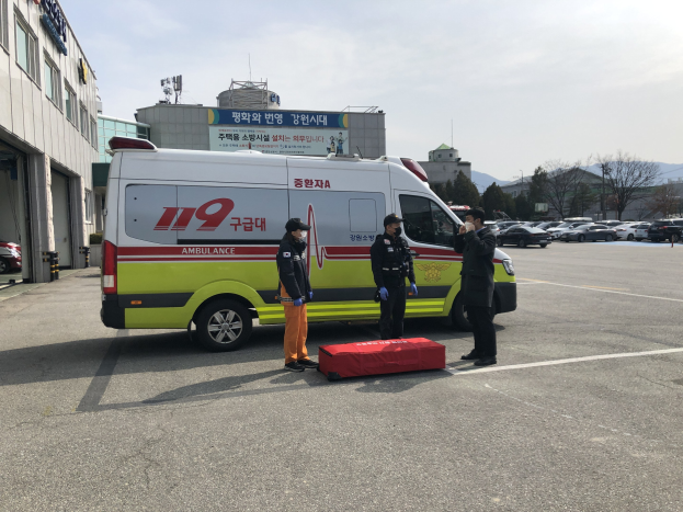 Ambulanz auf einem Parkplatz neben einem gl├Ąserfrontigen Geb├Ąude geparkt, mit drei Personen auf der Stra├če davor, umgeben von anderen Fahrzeugen, B├Ąumen, Bergen und Himmel.