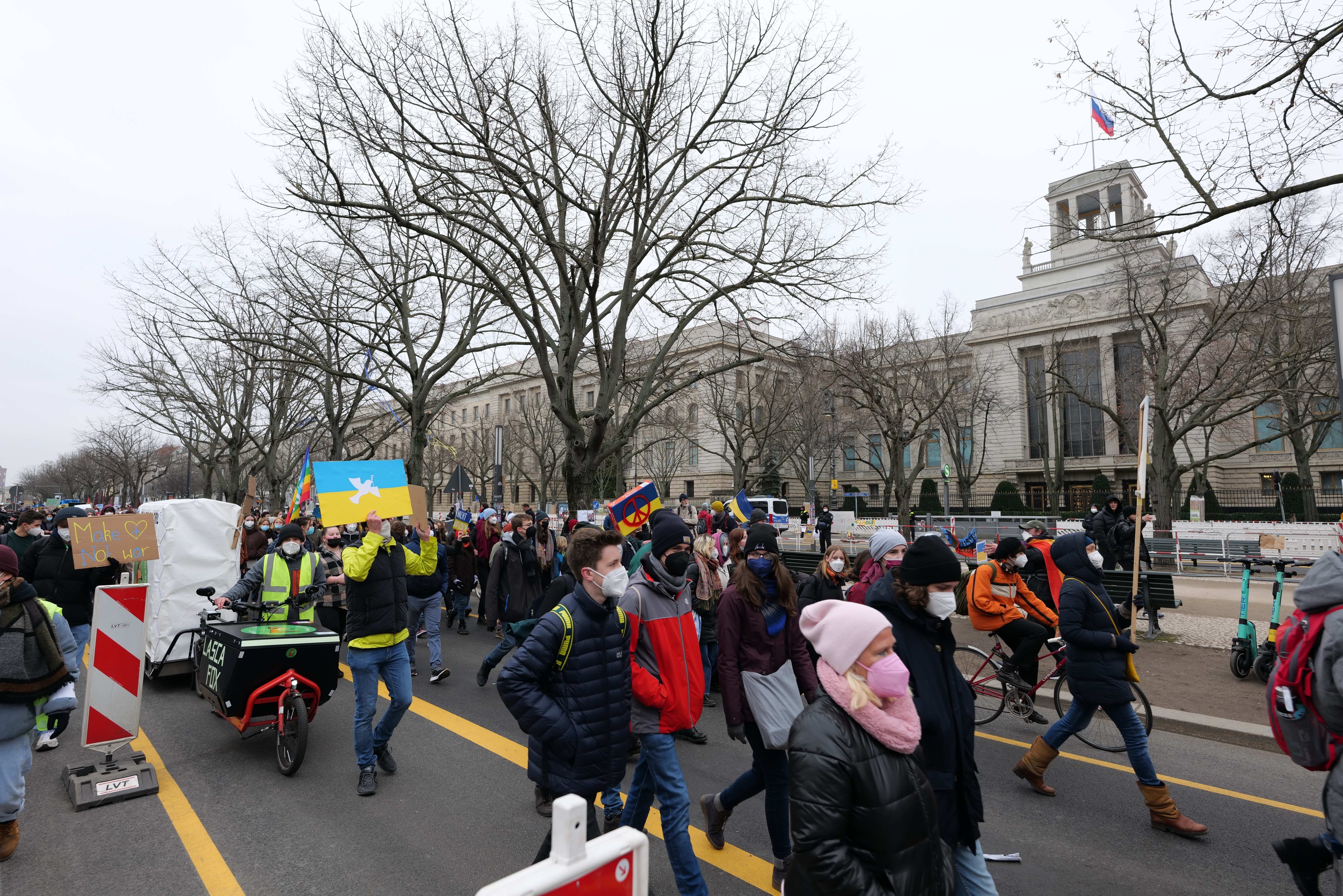 Eine große Gruppe von Menschen nimmt an einer Protestmarsch auf einer Straße in Washington, D.C. teil, mit Schildern und Transparenten, einige fahren Fahrräder, mit Bäumen, Schildern und einem klaren blauen Himmel im Hintergrund.