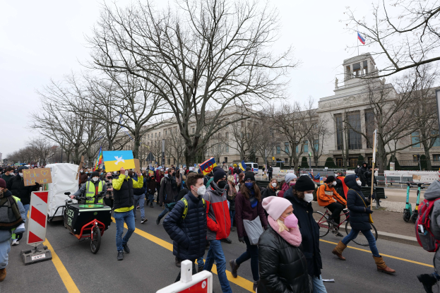 Eine große Gruppe von Menschen nimmt an einer Protestmarsch auf einer Straße in Washington, D.C. teil, mit Schildern und Transparenten, einige fahren Fahrräder, mit Bäumen, Schildern und einem klaren blauen Himmel im Hintergrund.