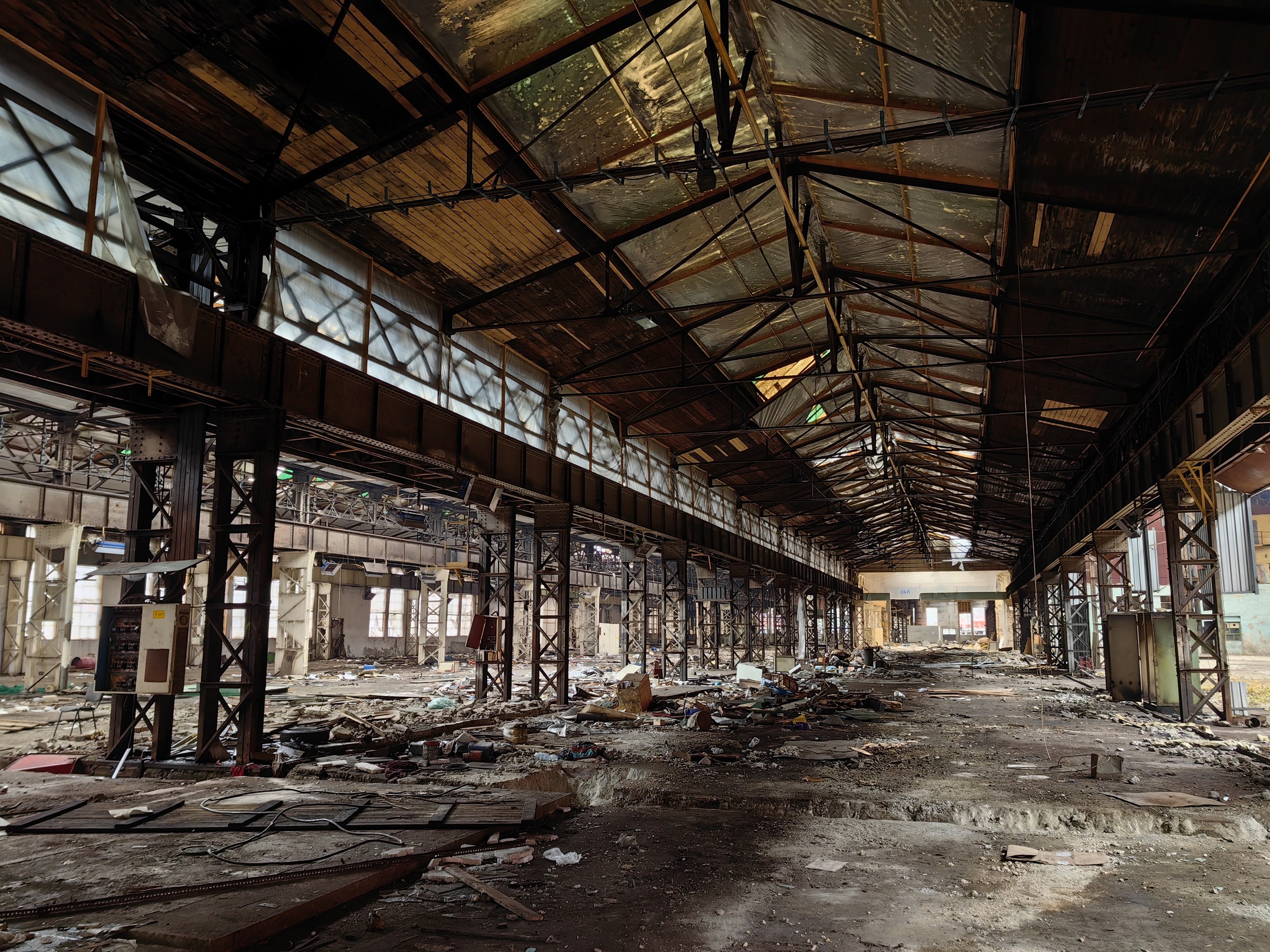 Verlassene Fabrikhalle mit verstreuter Trümmern, kaputten Fenstern, Metallstangen, Deckenlampen und einem beschädigten Dach.