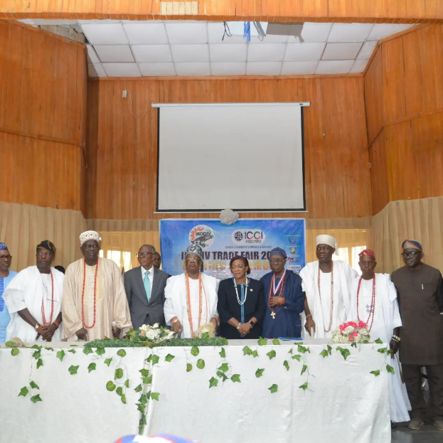 Eine Gruppe von Menschen, die um einen mit einem weißen Tuch bedeckten Tisch stehen, der mit Blumen und Blättern geschmückt ist, mit einem Banner im Hintergrund mit dem Text "ICCI International Trade Fair 2019", einem Projektionsschirm und Deckenleuchten.