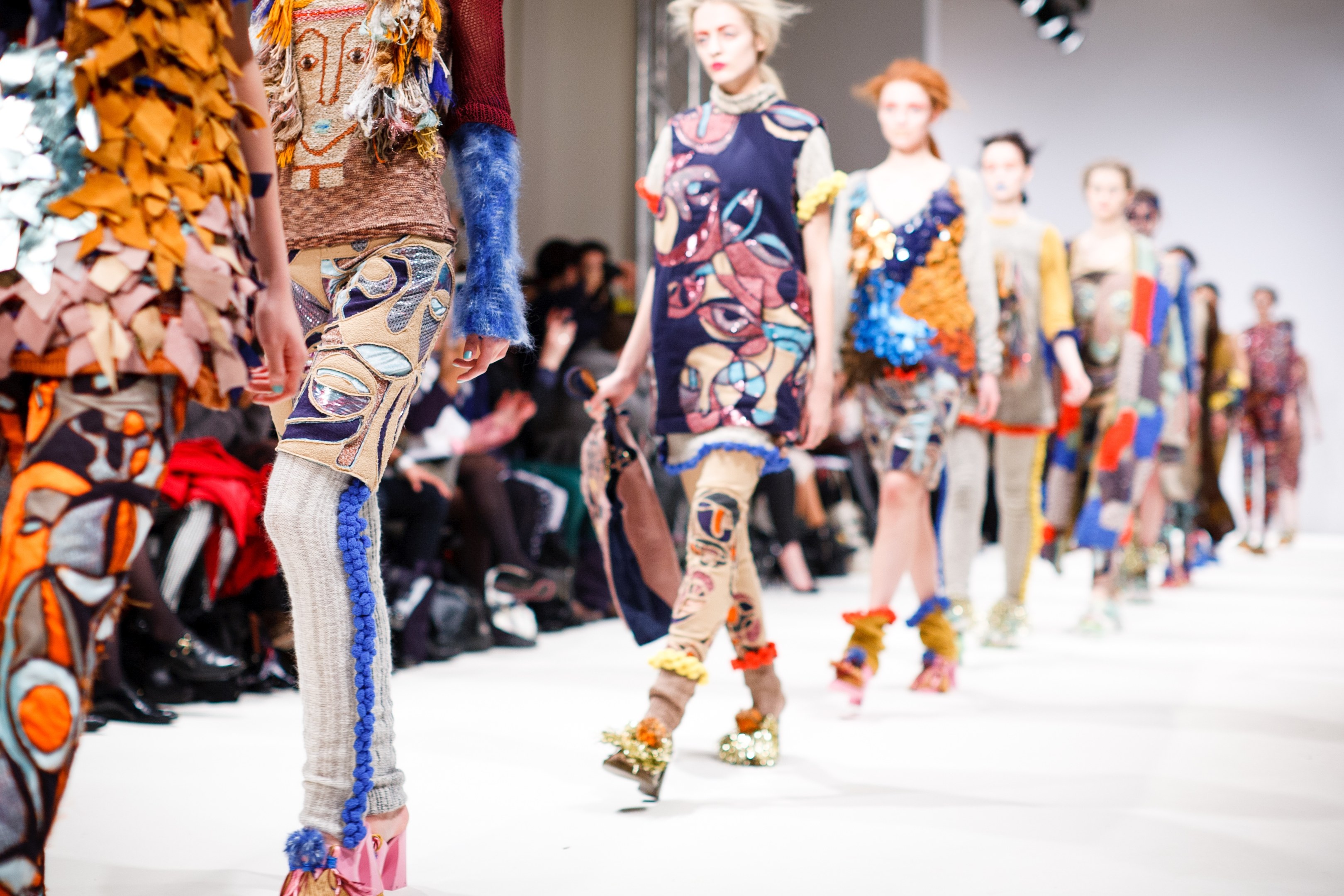 Eine Gruppe von Frauen, die auf dem Laufsteg der London Fashion Week gehen, mit einem Publikum im Hintergrund unter hellen Scheinwerfern.