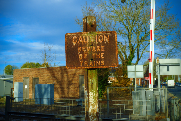 Warnschild an einem Bahnölzaun neben Bäumen, Masten, einem Gebäude, Containern, Fahrzeugen und einem bewölktem Himmel.