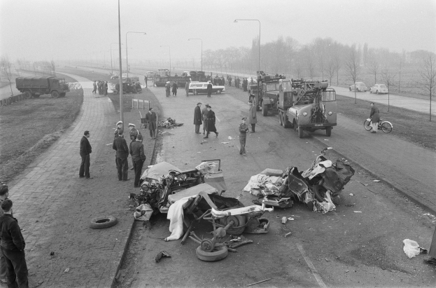 Schwarz-weiß-Bild eines Autounfalls am Straßenrand mit mehreren Fahrzeugen und einer Gruppe von Menschen in der Nähe, Laternenpfähle, Bäume und Himmel im Hintergrund.