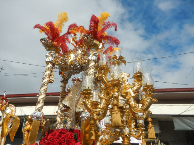 Ein großes goldenes und rotes Festwagen, geschmückt mit Blumen und anderen Dekorationen, bei einem Karnevalsumzug, mit einem Gebäude, Strommasten mit Drähten und einem bewölkten Himmel im Hintergrund.