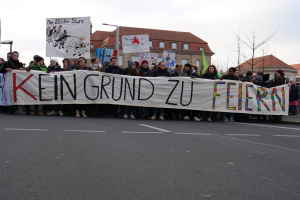 Demonstranten mit einem Transparent mit der Aufschrift 'Kein Grund zu Feiern' gegen deutsche Sparmaßnahmen, mit Straßeninfrastruktur und Gebäuden im Hintergrund bei klarem Himmel.