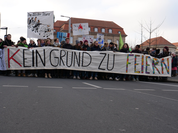 Demonstranten mit einem Transparent mit der Aufschrift 'Kein Grund zu Feiern' gegen deutsche Sparmaßnahmen, mit Straßeninfrastruktur und Gebäuden im Hintergrund bei klarem Himmel.