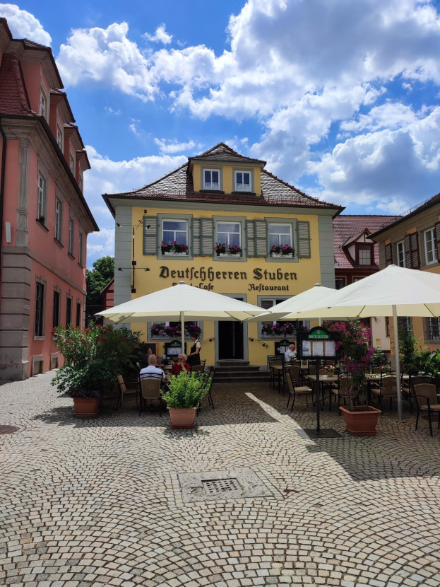Außensitzbereich mit Tischen und Sonnenschirmen vor einem gelben Gebäude mit der Aufschrift "Deutschen Stuben Restaurant" in Baden-Württemberg, Deutschland, mit einigen Menschen, die sitzen und stehen, unter einem blauen und weißen Himmel.