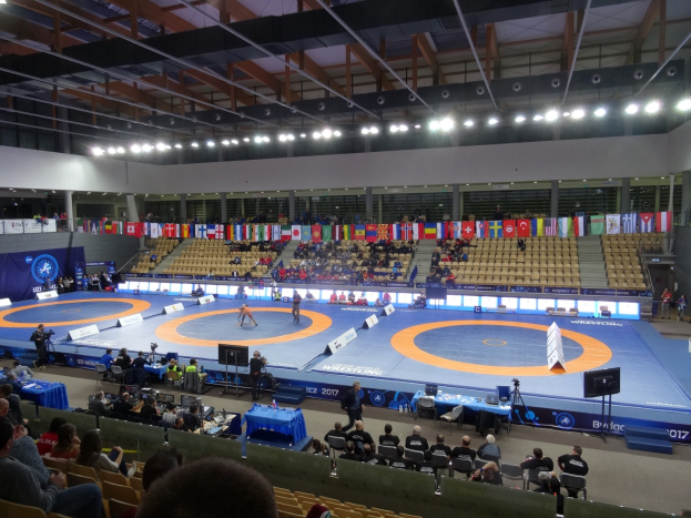 Großer Hallenkampf mit Zuschauern bei einem Wrestling-Match, einschließlich sitzender und stehender Zuschauer, Tischen mit Gegenständen und Hintergrundflaggen, Lichtern und Stangen.