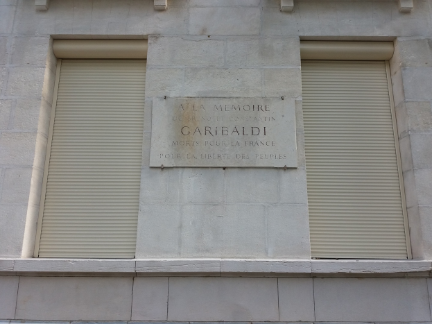 Ein Gebäude mit zwei Fenstern und Läden sowie einer Gedenktafel an der Seite.