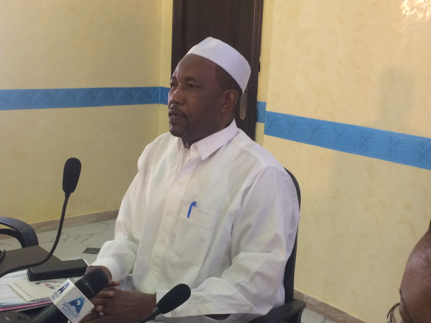 Ein Mann in einem weißen Gewand und Hut sitzt an einem Tisch mit Mikrofonen, spricht, während Papiere und Gegenstände auf dem Tisch liegen, mit einer Person mit Brille rechts von ihm, in einem Raum mit einer Wand und einer Tür im Hintergrund.