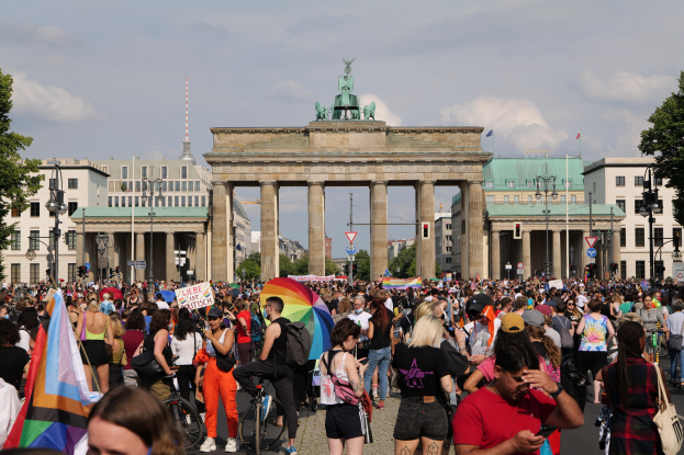 Eine große Menschenmenge marschiert vor dem Brandenburger Tor in Berlin, Deutschland, vorbei, carrying flags and banners, with some on bicycles; the street is lined with light poles and trees under a cloudy sky, and buildings with windows and arches, including a statue, are visible in the background.