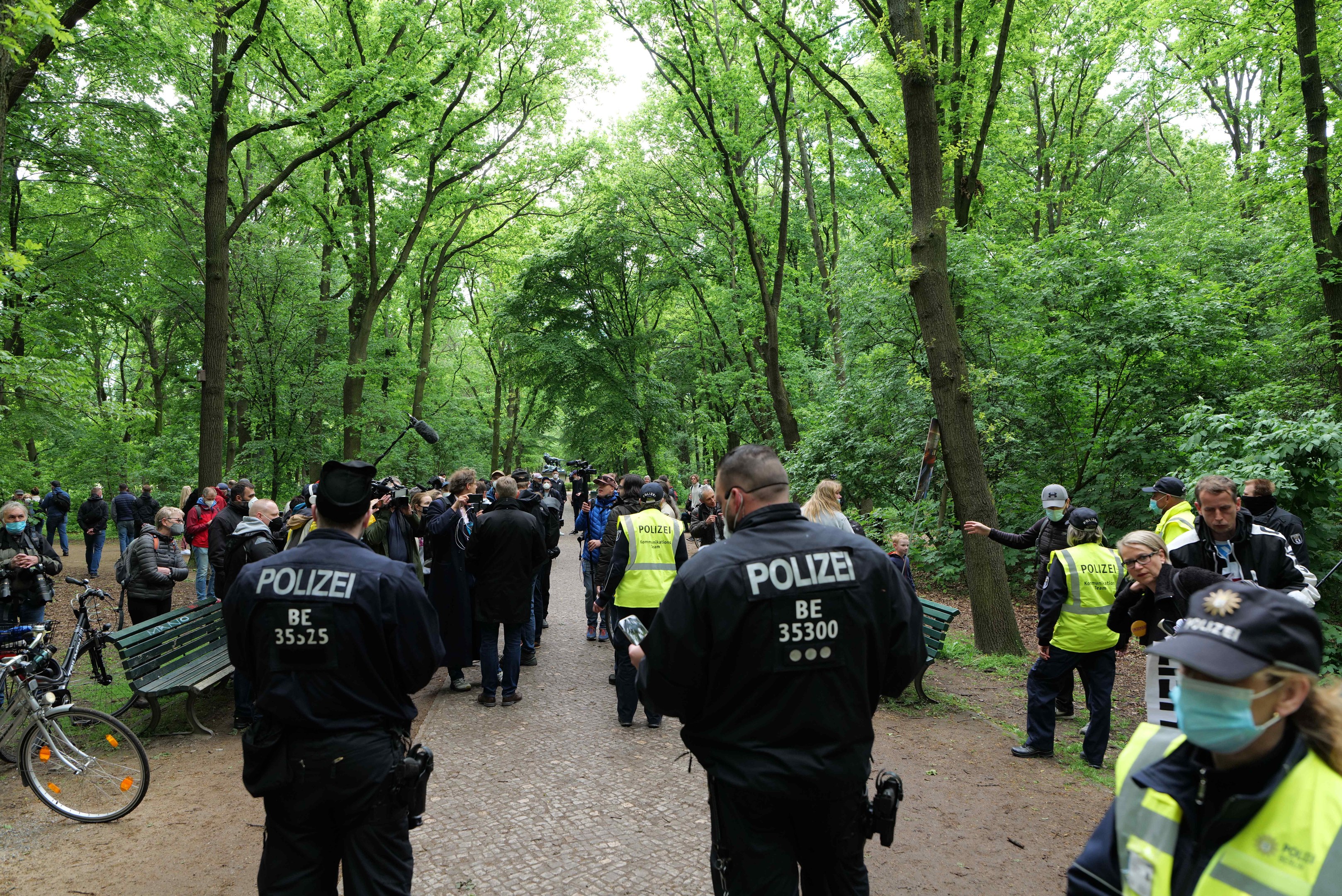Polizisten vor einer Menschenmenge bei einer Outdoor-Veranstaltung, mit Fahrrädern und einer Bank im Vordergrund und Bäumen im Hintergrund.