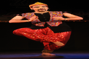 Ein Mann in roter und schwarzer Kleidung tanzt auf einer Bühne mit dunklem Hintergrund, seine Augen sind geschlossen und sein Mund leicht geöffnet.