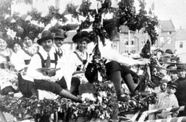 Schwarzes und weißes Bild eines Paradewagens mit Menschen in traditioneller bayrischer Tracht, einige spielen Instrumente, mit Blattdekoration vor einem Hintergrund von Gebäuden unter einem klaren Himmel.