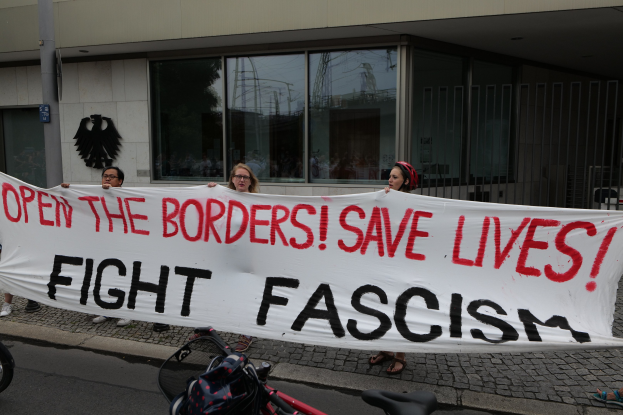Gruppe von Menschen mit einem Banner "Öffnet die Grenzen, rettet Leben, kämpft gegen Faschismus" vor einem Gebäude mit Glasfenstern und einem Pfahl, mit einem Fahrrad und einer Tasche auf der Straße.