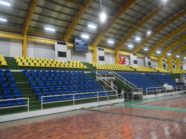 Großer Hallensportplatz mit Holzboden, blauen und gelben Sitzplätzen, Geländern, Stufen zum Sitzbereich, wandmontierten Bildschirmen, Deckenleuchten und einigen Menschen.