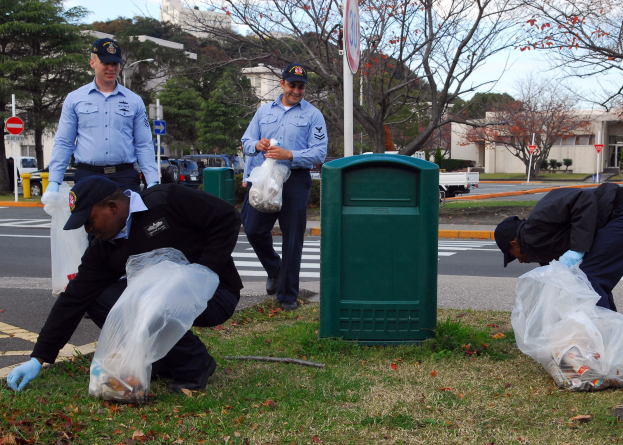 Drei Polizisten in Uniform sammeln Müll am Straßenrand, mit Bäumen, Gebäuden, Fahrzeugen und einem Abfalleimer im Hintergrund.