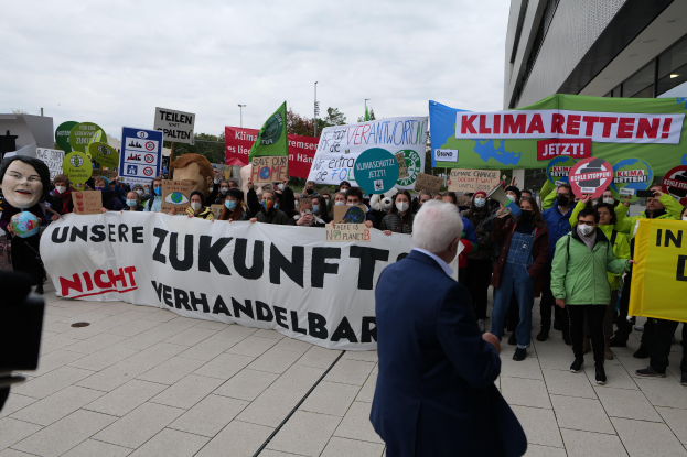 Eine Gruppe von Menschen hält Schilder und Transparente und protestiert gegen den Klimawandel vor einem Gebäude in Deutschland, wobei eine Person auf der linken Seite eine Kamera hält, Bäume, Pfähle und einen klaren blauen Himmel im Hintergrund zu sehen sind.
