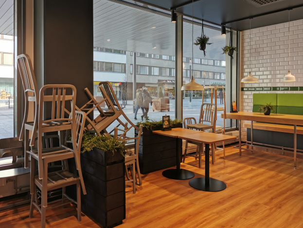 Ein Restaurantinnenraum mit Holztischen und -stühlen, großen Fenstern, die eine Stadtstraße mit Menschen, Gebäuden und Himmel zeigen, umgeben von Pflanzen und Lichtern.