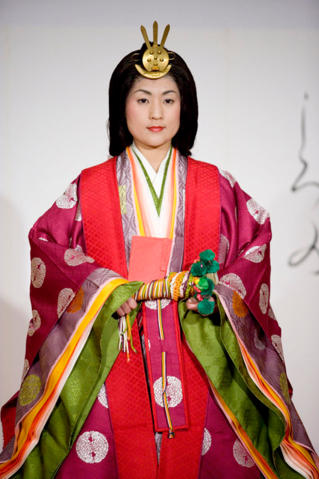 Eine Frau in einem traditionellen japanischen Kimono, das ein leuchtendes rotes und grünes Kleid mit aufwändigen Mustern zeigt, steht selbstbewusst auf einem Laufsteg mit ihrem Haar zu einem ordentlichen Knoten gebunden.